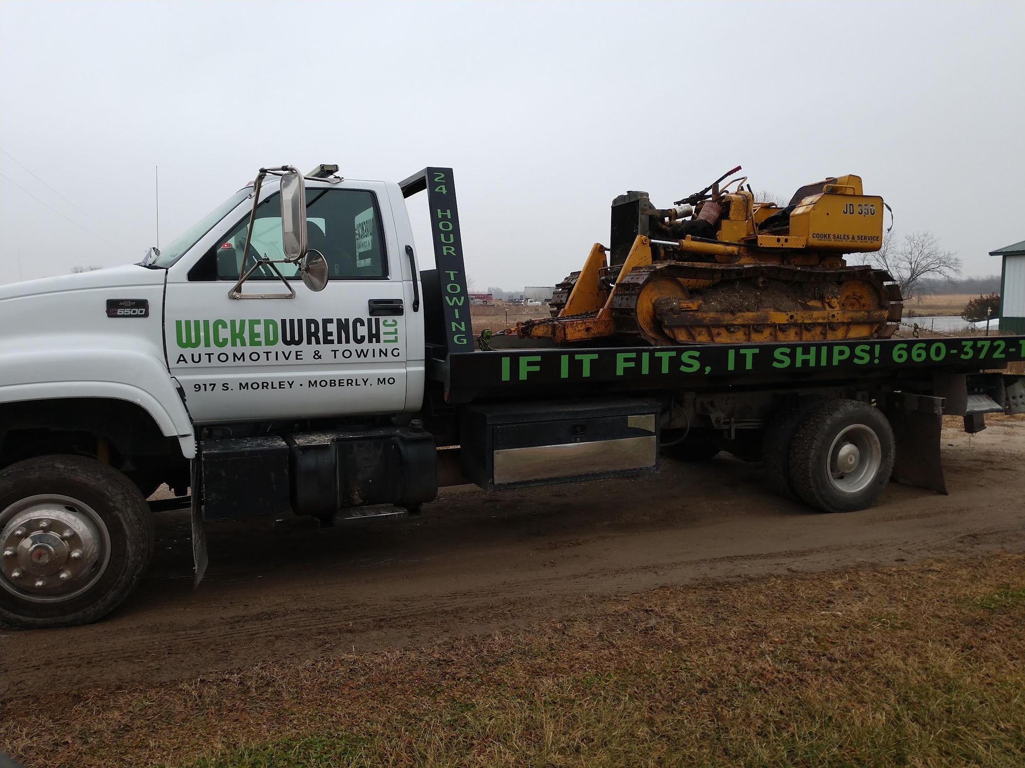 Wicked Wrench flatbed truck hauling a John Deere dozer, lettered with 24 Hour Towing and If It Fits, It Ships